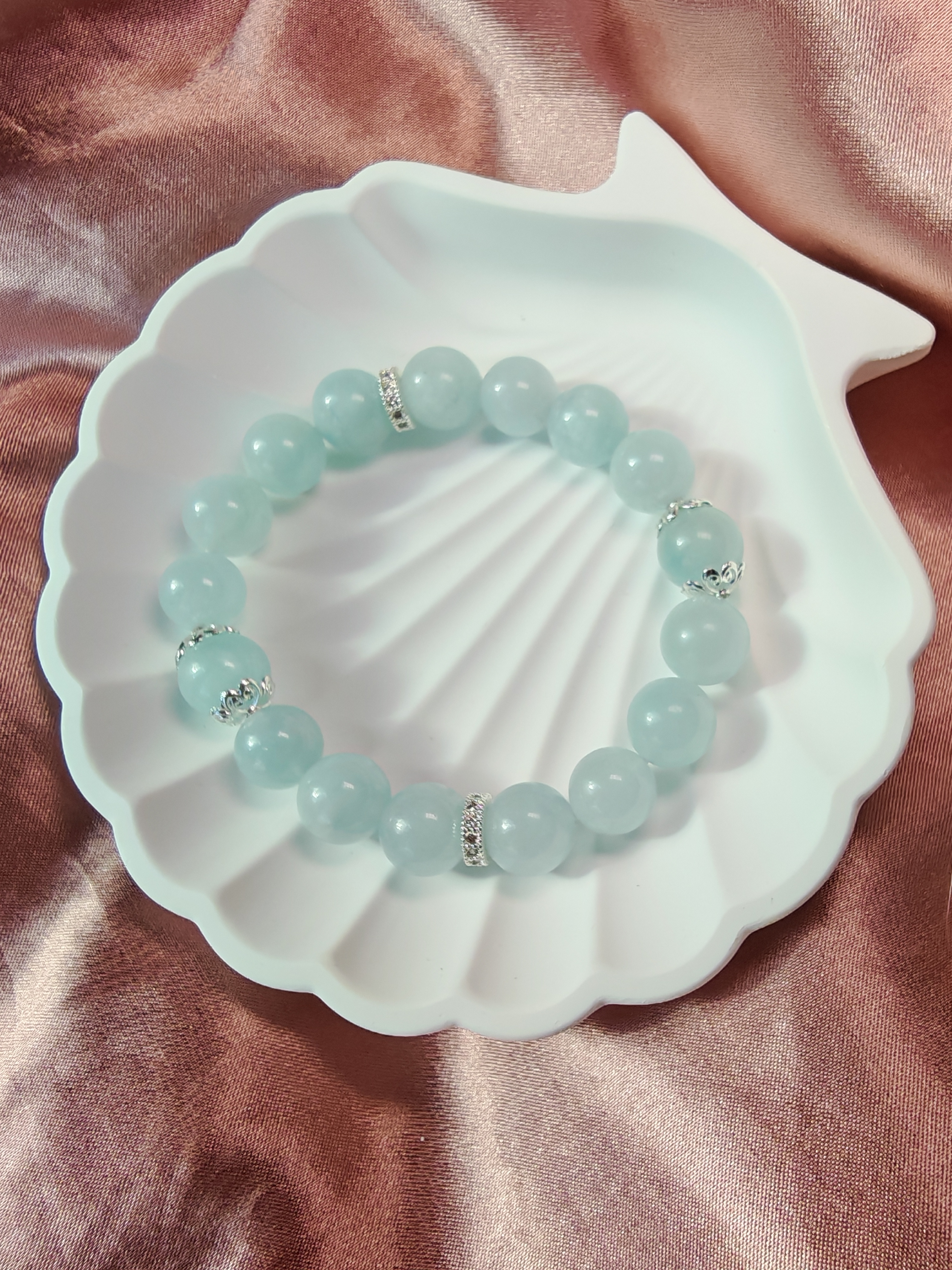 Brățară din mărgele de acvamarin bleu deschis cu distanțiere argintii cu cristale, așezată într-un suport alb în formă de scoică.